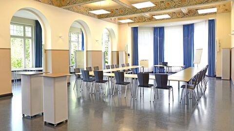 Aula Innenansicht mit vielen Fenstern, Tischen und Stühlen