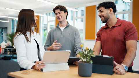 Eine junge Frau und zwei junge Männer stehen an einem Stehtisch in einem Raum des Campus Frankfurt am Main.